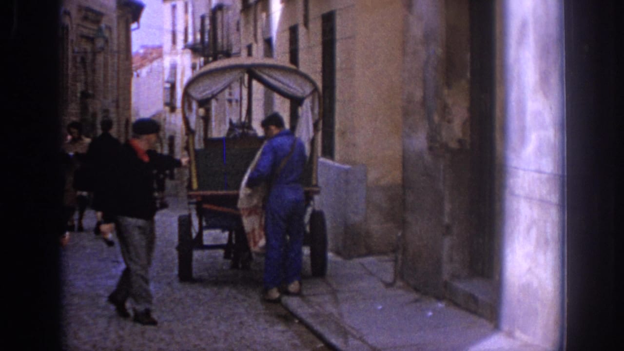 1966 Spain street scene with pedestrians, a covered cart, and a worker in blue beside stone buildings.
