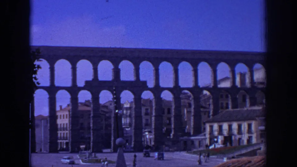 Static 1966 home-movie frame of the Aqueduct of Segovia above a square with pedestrians, cars, and multi-story buildings.