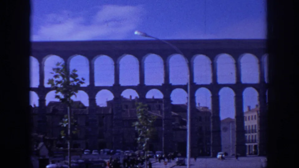 1966 Spain street-level view of the aqueduct with a streetlamp, small trees, cars, pedestrians, and surrounding buildings.