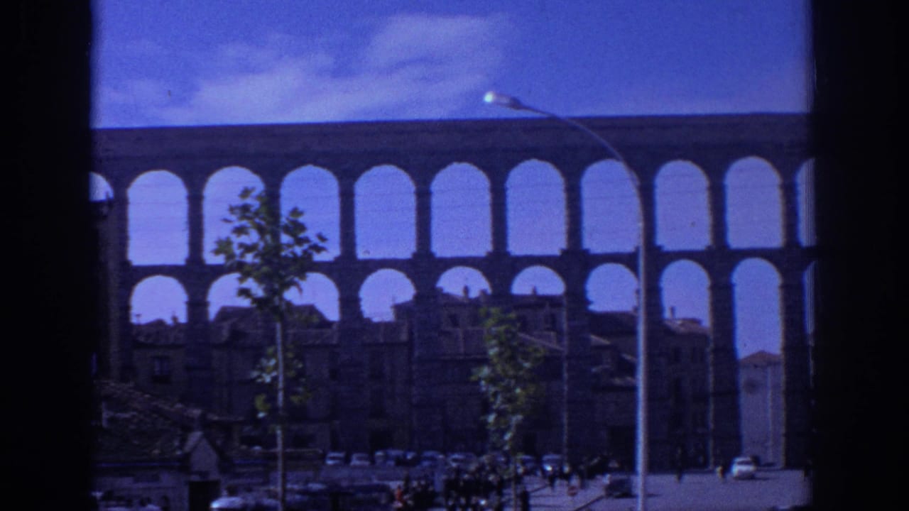 1966 Spain home-movie view of the Aqueduct of Segovia through a dark window frame, with rooftops, trees, cars, and tiny pedestrians below.