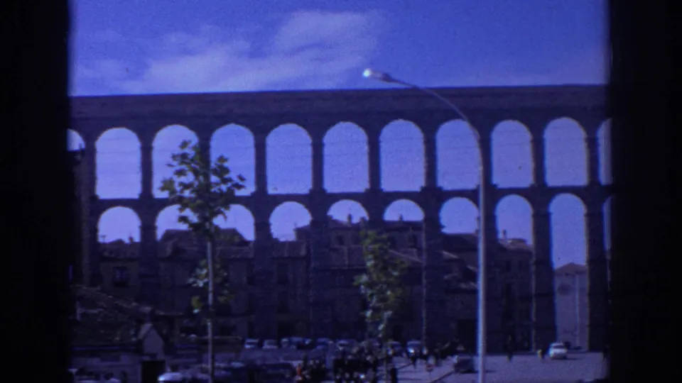 1966 Spain home-movie view of the Aqueduct of Segovia through a dark window frame, with rooftops, trees, cars, and tiny pedestrians below.