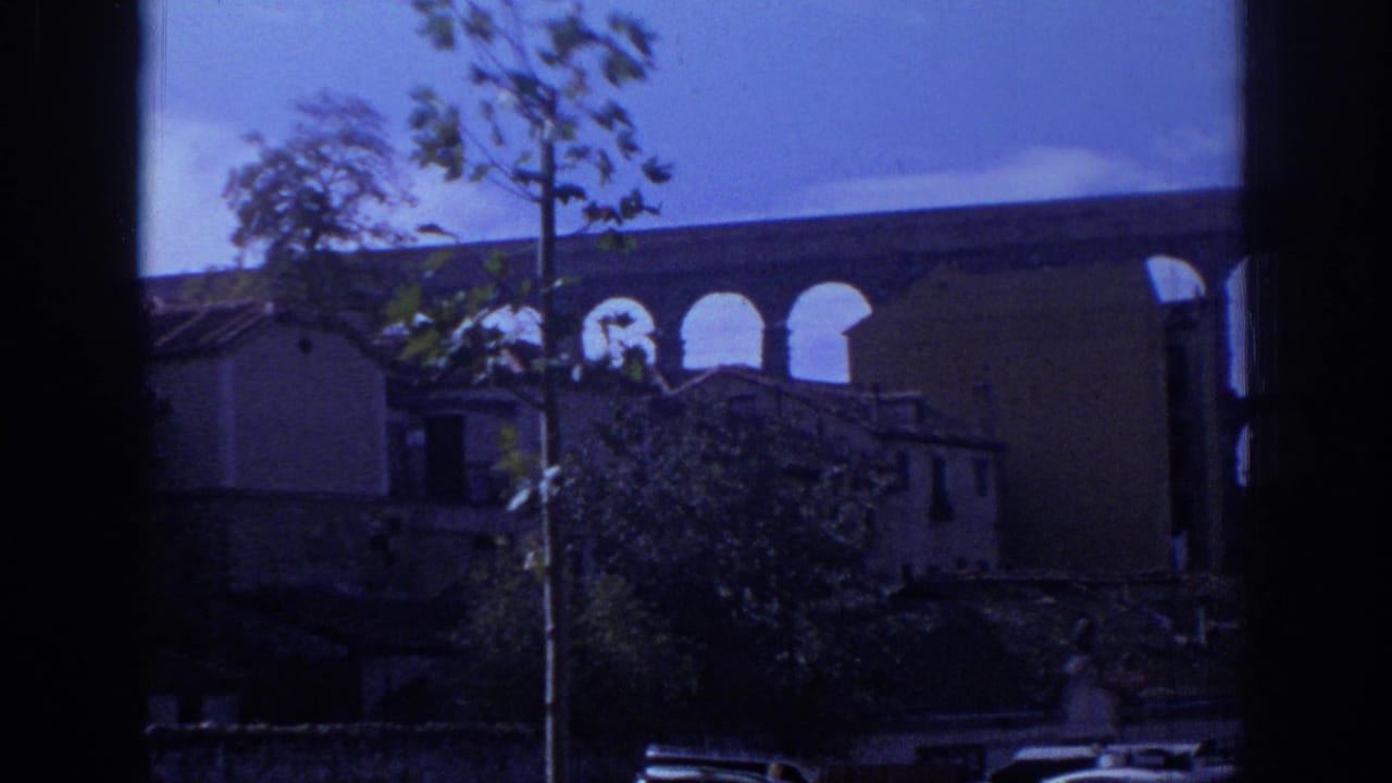 Blue-tinted 8mm view of the Segovia aqueduct partly hidden by nearby buildings, trees, parked cars, and a dark window or vehicle frame.