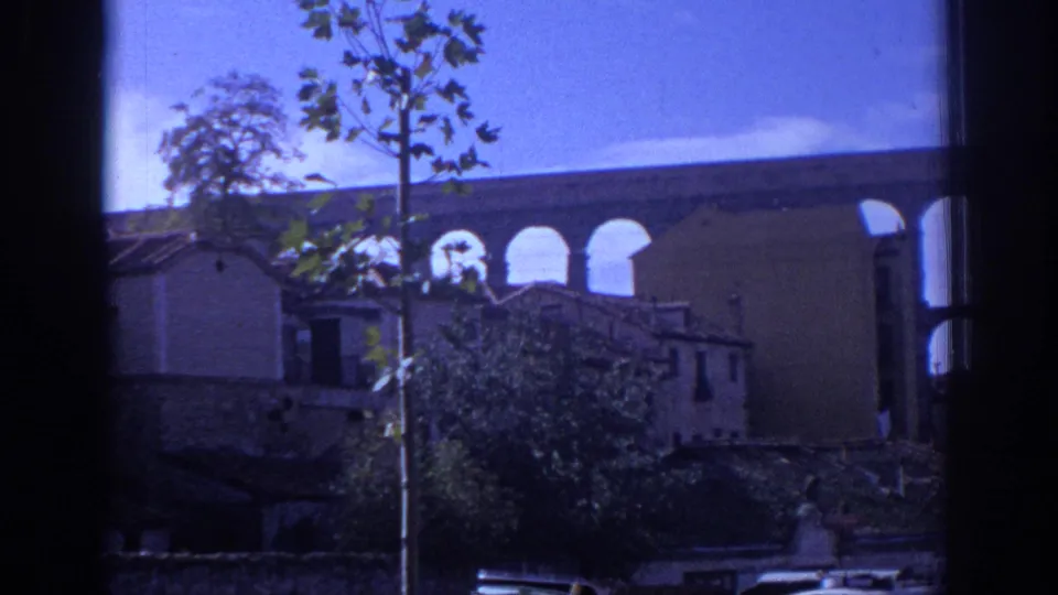 1966 Segovia travel-film frame showing trees, low buildings, and portions of the aqueduct behind them, with cars along the lower street edge.