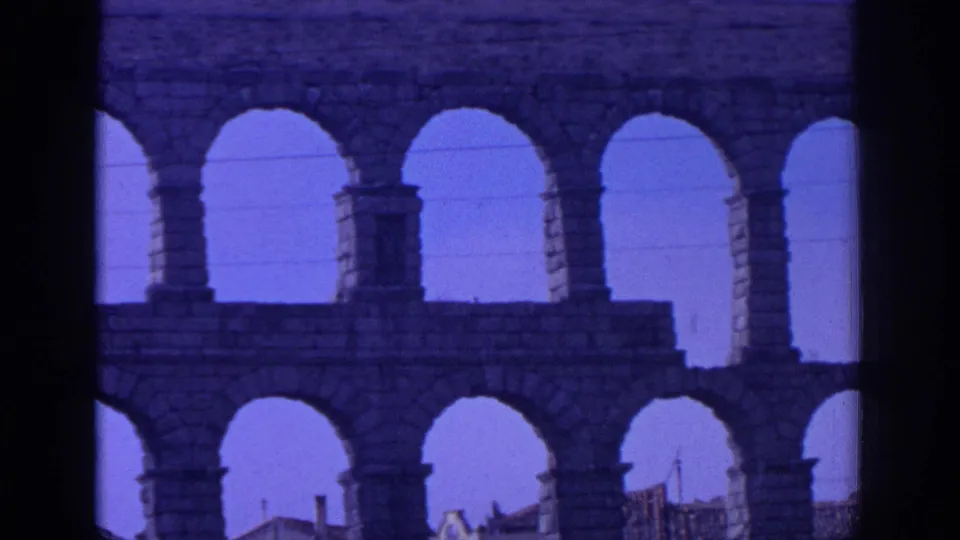 Closer 8mm frame of the Aqueduct of Segovia, emphasizing the stone piers, upper and lower arches, bright sky, and rooftops below.