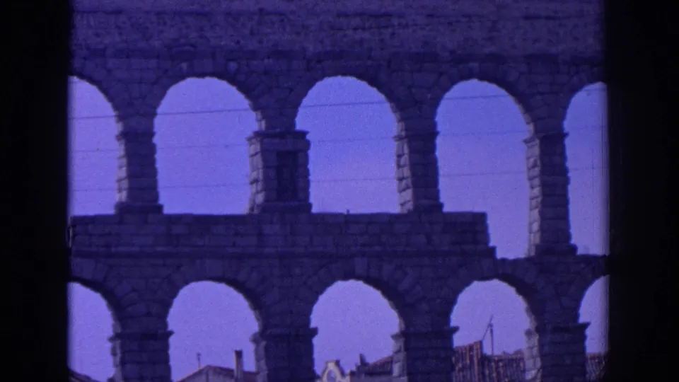 1966 Spain 8mm view through the aqueduct, showing repeated stone arches, tall piers and rooftops behind them.
