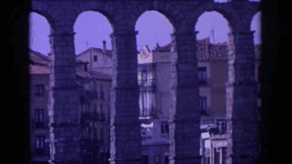 1966 Spain 8mm frame showing the aqueduct's lower piers with Segovia buildings, tiled roofs and street area behind.