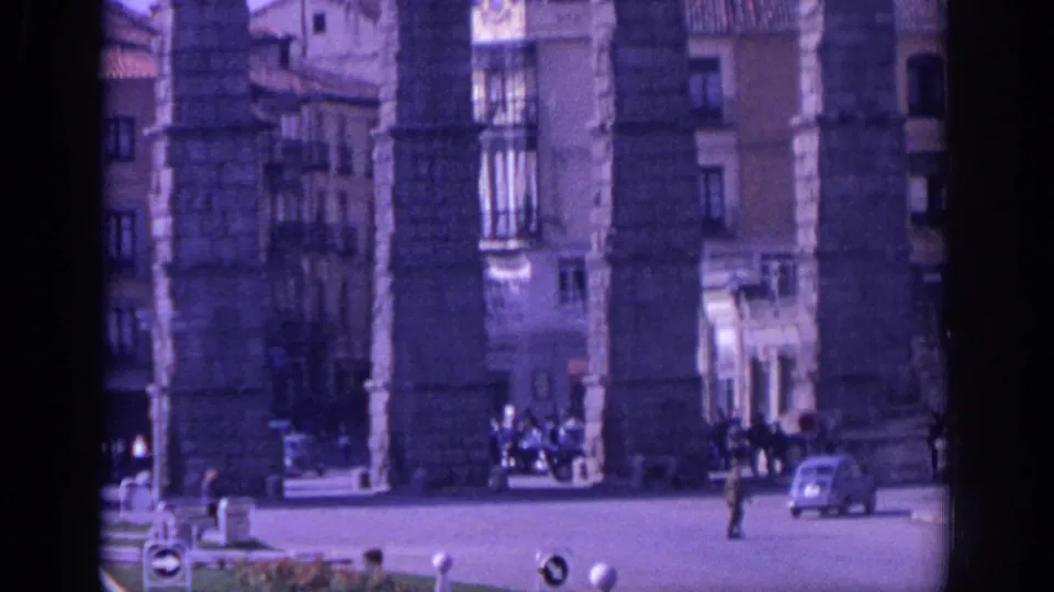 1966 Spain home-movie view of massive aqueduct piers, nearby buildings, pedestrians, and small period cars in a city square.