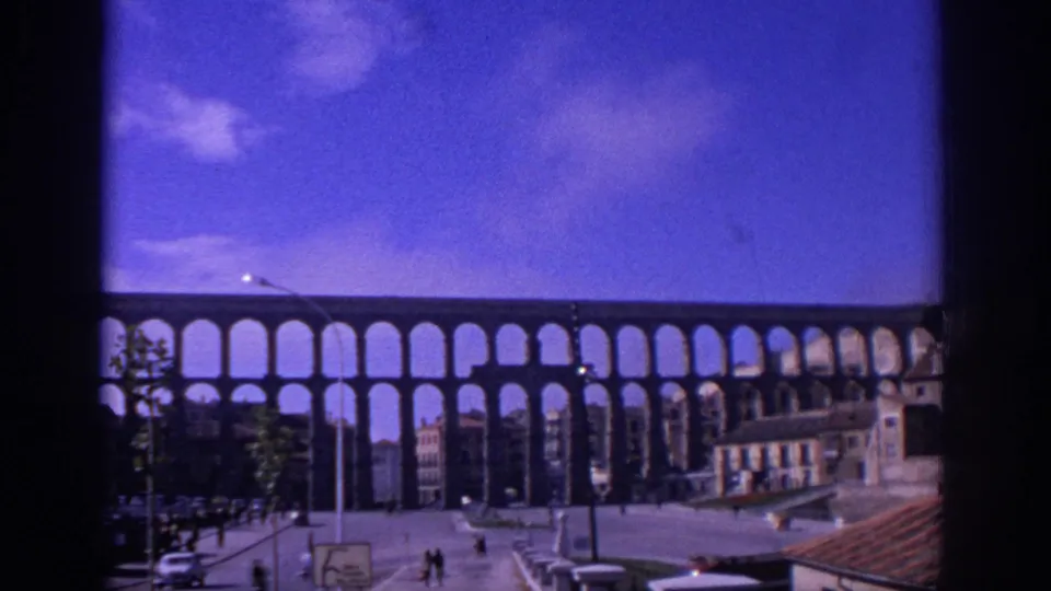 Wide 1966 Spain view of the Segovia aqueduct crossing the frame, with pedestrians, cars, rooftops, and lamp posts below.