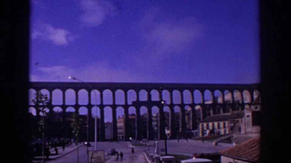 1966 Spain travel-film frame showing the Segovia aqueduct above a plaza with small figures, period cars, street signs, and nearby buildings.