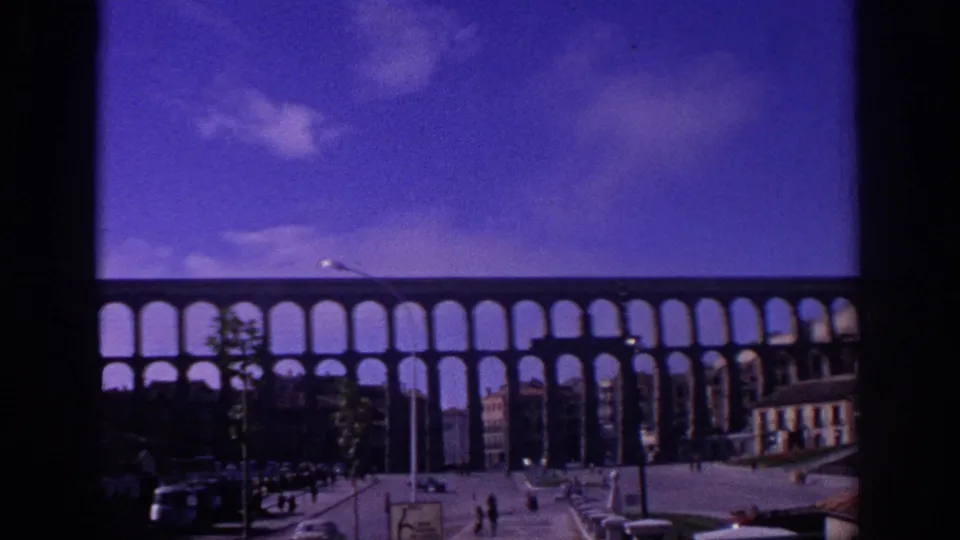 Soft 8mm frame from 1966 Spain with Segovia's aqueduct, a broad blue sky, pedestrians, cars, lamp posts, and tiled rooftops.