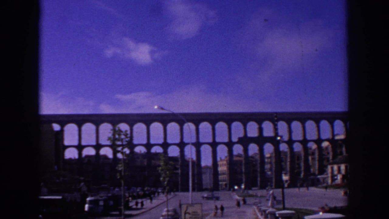 1966 Segovia street view of the Roman aqueduct with repeated arches, cars, pedestrians, young trees, and color-shifted 8mm sky.