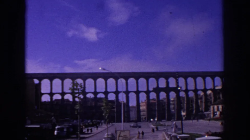 1966 Segovia street view of the Roman aqueduct with repeated arches, cars, pedestrians, young trees, and color-shifted 8mm sky.