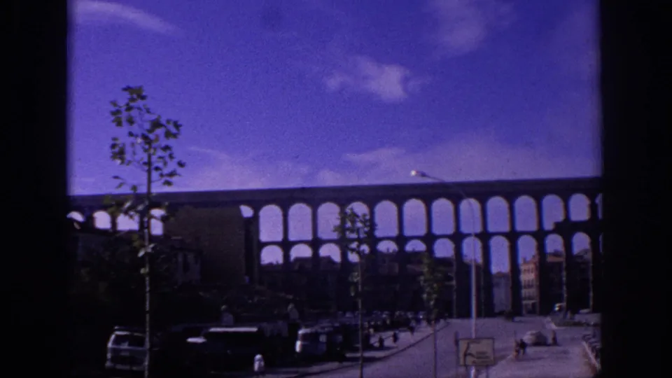 Color-shifted 8mm view of the Segovia aqueduct behind young trees, cars, pedestrians, and dark foreground street shadows.