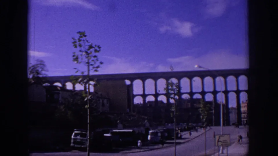 1966 Segovia street scene with the Roman aqueduct, young trees, cars, small pedestrians, buildings, and strong 8mm color shift.
