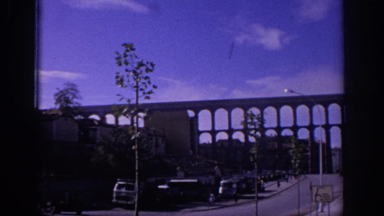 1966 Spain home-movie view of a multi-arched aqueduct or bridge, street traffic, young trees, and a small road sign under a purple-blue sky.