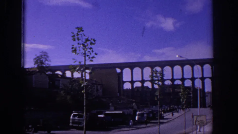 1966 Spain home-movie view of a multi-arched aqueduct or bridge, street traffic, young trees, and a small road sign under a purple-blue sky.