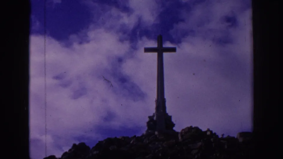 A tall cross monument in Spain appears on a rocky hillside beneath moving clouds, with visible 8mm grain and purple-blue color shift.