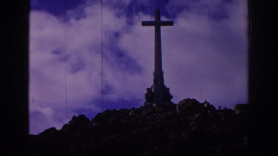 1966 Spain home-movie frame of a tall hilltop cross, rocky foreground, clouds, and a dark window or film-frame border at the side.