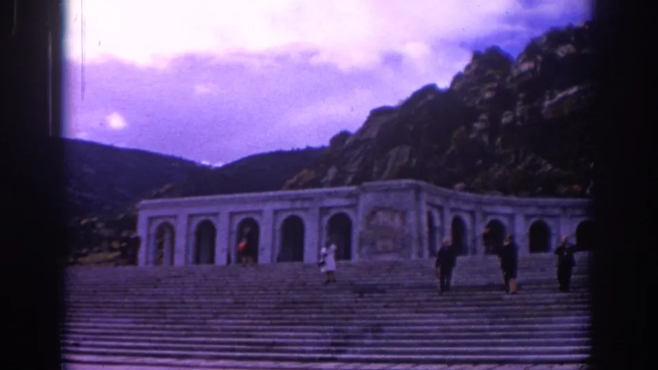 1966 Spain home-movie frame with visitors scattered across monumental stairs before a stone arcade and mountain backdrop.