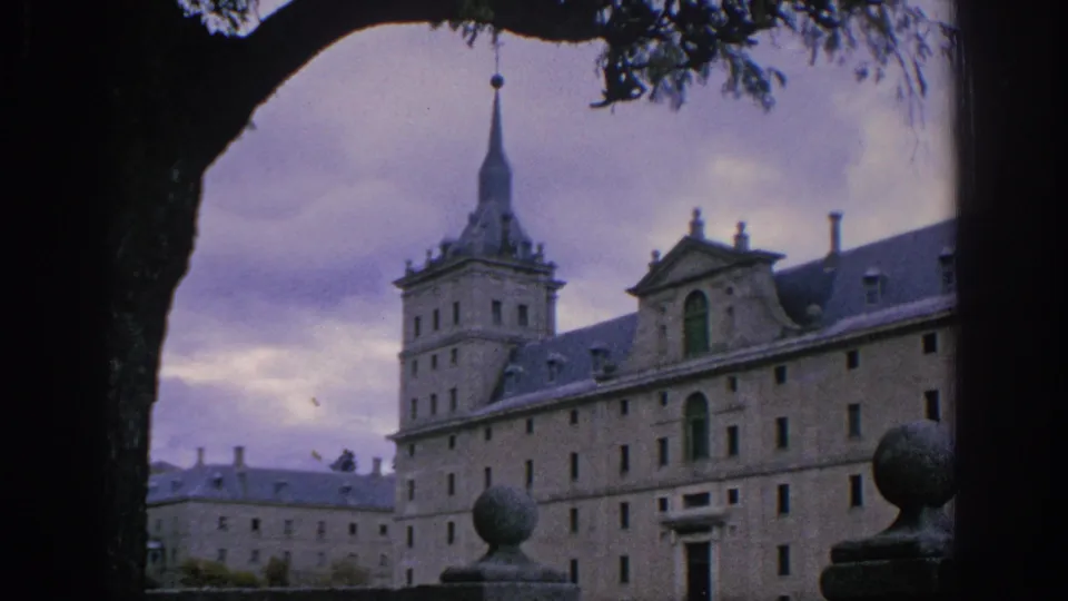 1966 Spain travel frame showing a long historic stone building, tower, spire, and balustrade ornament framed by a tree.