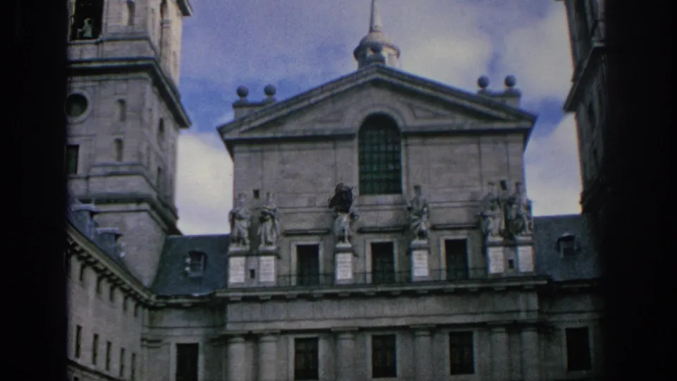 1966 Spain 8mm frame showing a closer view of a historic stone facade with tower, pediment, arched window, statues, and columns.
