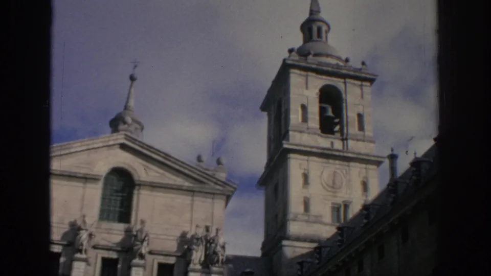 1966 Spain 8mm frame looking up at a bell tower and historic stone facade with visible roofline statues and arched window detail.