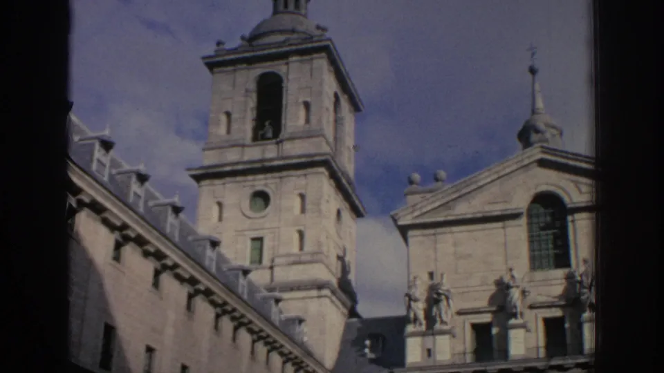 1966 Spain 8mm frame showing a bell tower, roofline statues, slate-like roof sections, and a historic stone facade in a courtyard view.