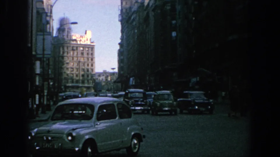 1966 Spain street view showing mid-century cars traveling along a cobblestone urban road lined with tall buildings.