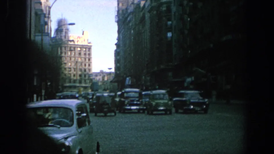 1966 Spain frame showing a light compact car in the foreground and other mid-century vehicles on a shadowed city street.