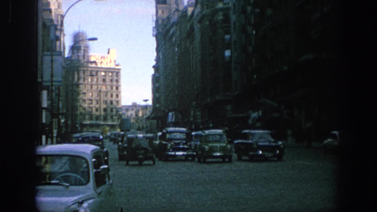Busy city street with compact cars, tall buildings, and a bright sky at the end of the avenue.