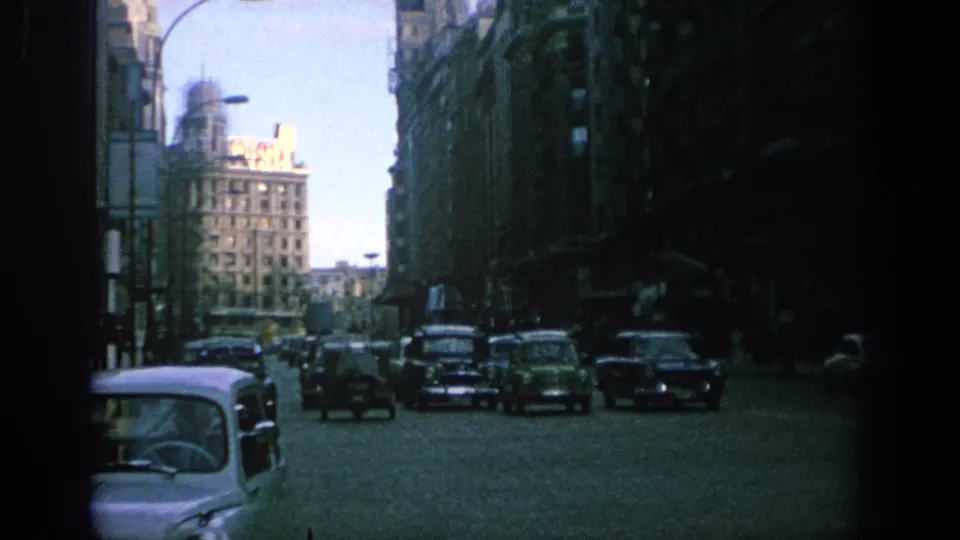 1966 Spain 8mm frame showing a broad city street with period cars, shaded building fronts, and a bright distant block.