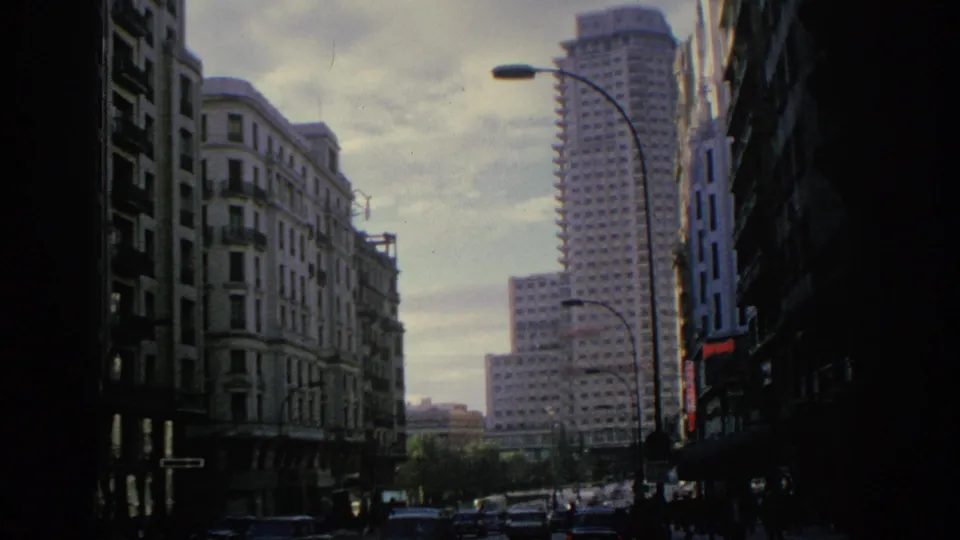 1966 Spain frame showing older urban facades, passing cars, and a prominent modern high-rise in the distance.