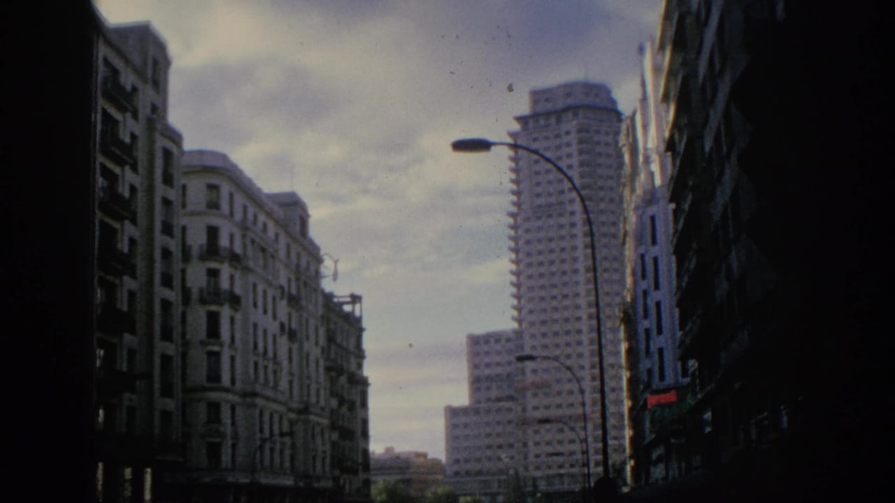1966 Spain home-movie frame showing a city street canyon with older apartment facades, curved lamps, and a prominent high-rise.