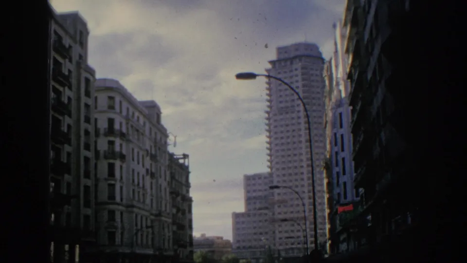 1966 Spain home-movie frame showing a city street canyon with older apartment facades, curved lamps, and a prominent high-rise.