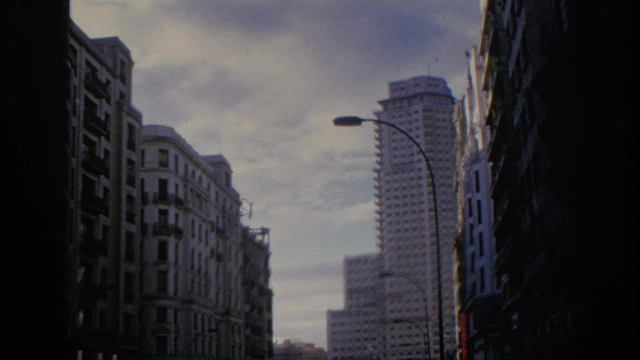 1966 Spain travel footage frame with apartment blocks, dark foreground facades, street lamps, and a distant modern tower.
