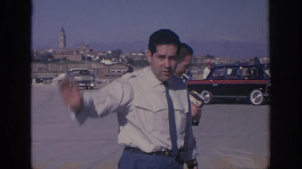 1966 Spain home-movie frame showing a man waving toward the camera beside 1960s cars, with a distant town and church tower.