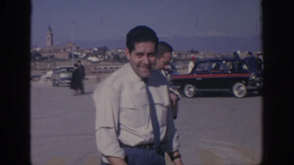 1966 Spain vacation-film frame showing a smiling man walking near classic cars with a church tower and town skyline behind him.