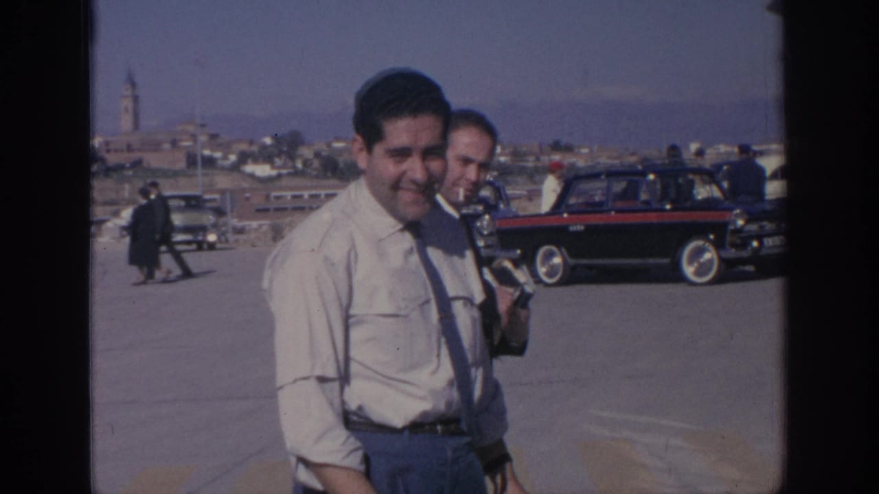 In Spain in 1966, a man in a light shirt and tie stands near a bus stop area with older cars and a distant town skyline.