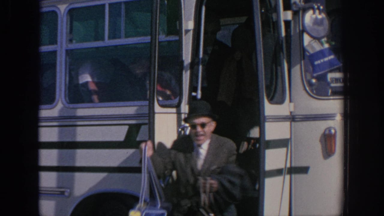 Older man in hat and sunglasses exits a white bus while carrying bags.