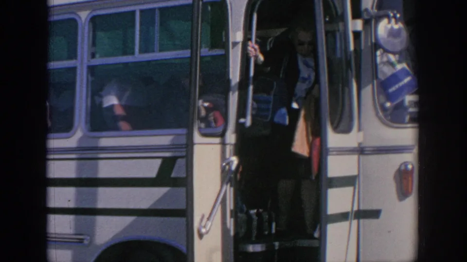 A traveler appears in the open doorway of the striped bus, partly in shadow, with bags visible near the steps.
