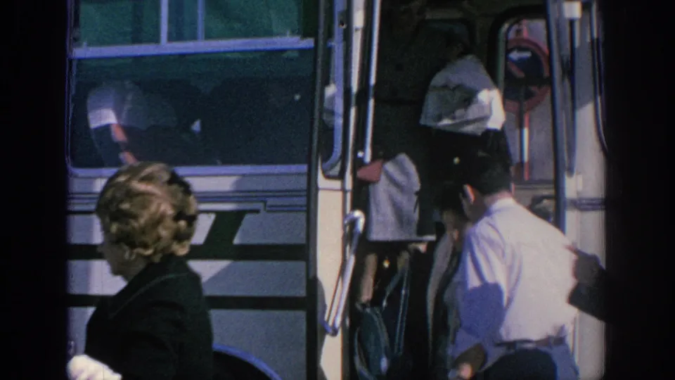 1966 Spain home-movie frame showing people beside a white tour bus, with a man in a light shirt assisting at the open door.