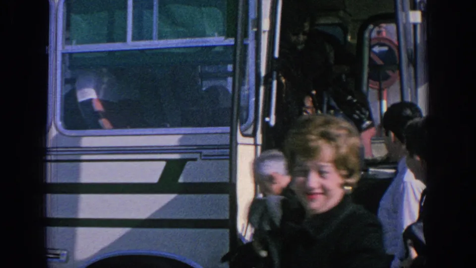 1966 Spain home-movie frame with a woman in a dark coat crossing foreground and travelers gathered around a white tour bus.