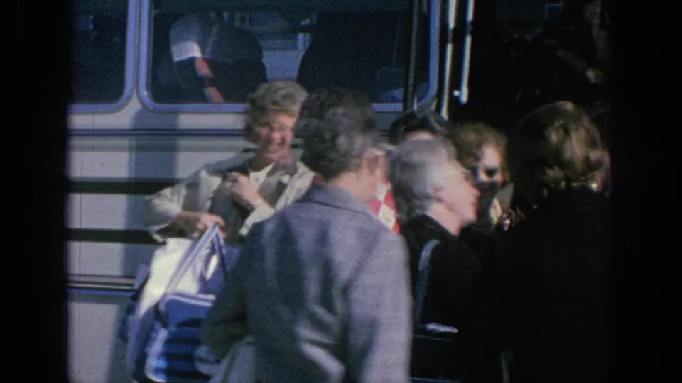 Adult tourists beside a white tour bus in Spain, with visible 1960s clothing, sunglasses, and travel bags.