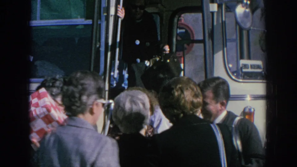 Adult tourists in 1960s attire gather beside a white tour bus during a Spain trip stop.