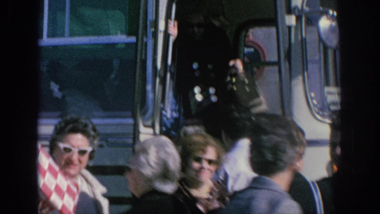 Spain 1966 home-movie frame showing adult travelers near a white tour bus, with sunglasses, coats, and a red patterned bag in view.