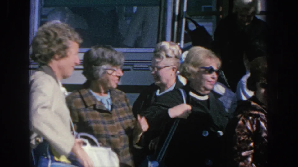 Spain 1966 home-movie frame with adult travelers clustered beside the bus, wearing glasses, patterned jackets, and light outerwear.
