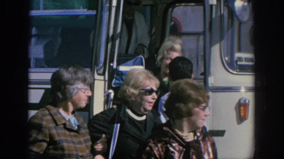 Spain 1966 frame showing adults moving away from an open tour bus door, with sunglasses, coats, and bus trim visible.