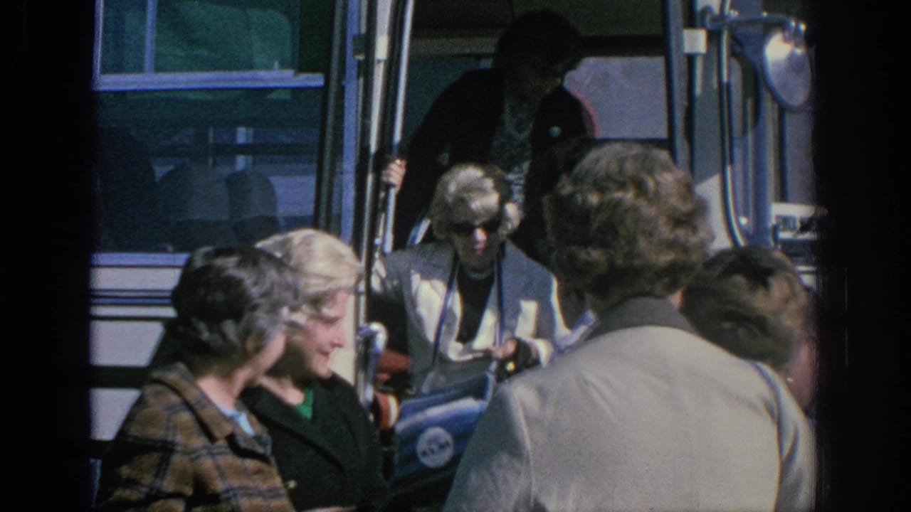 Older women in 1960s attire stand by a white tour bus in Spain, with one passenger carrying a blue NASA-branded bag.