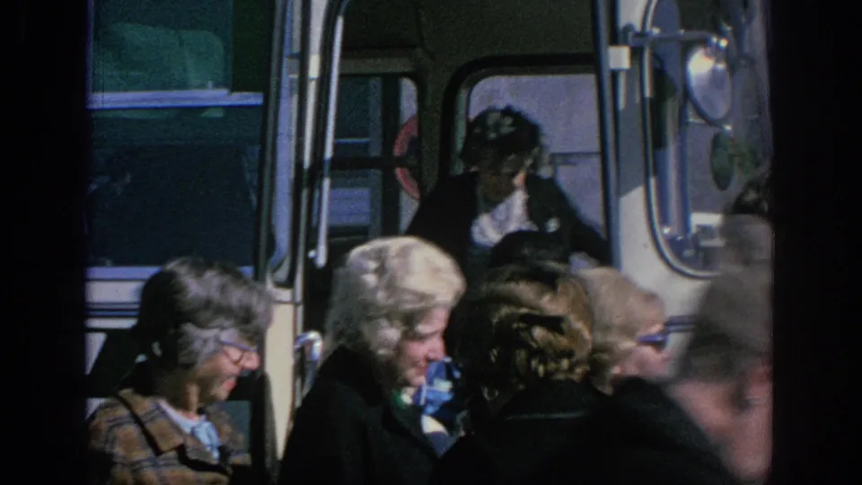 Crowded 1966 Spain travel scene with older women passing the open bus door and a blue NASA-branded bag visible among them.