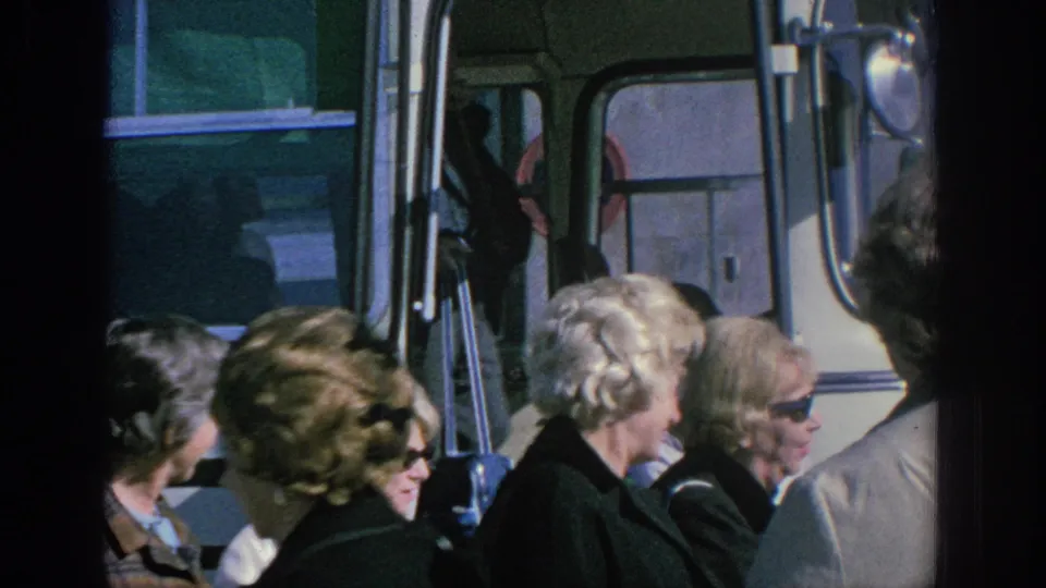 Older women in 1960s clothing walk beside a white tour bus in Spain, seen in soft 8mm color with the bus doorway behind them.